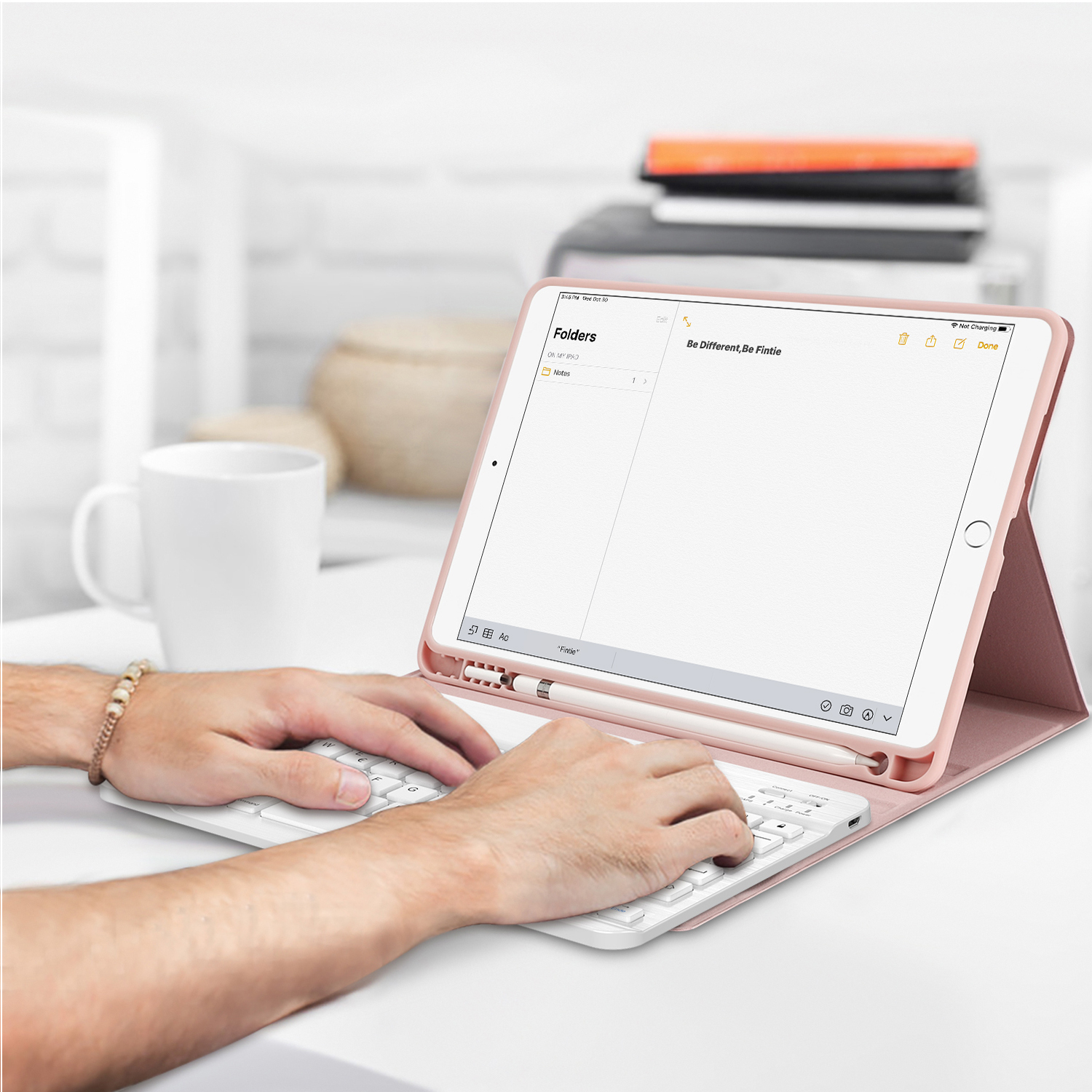 Hände, die auf einer weißen Tastatur tippen, neben einem geöffneten Tablet mit pinkem Gehäuse. Weiße Tasse sichtbar.