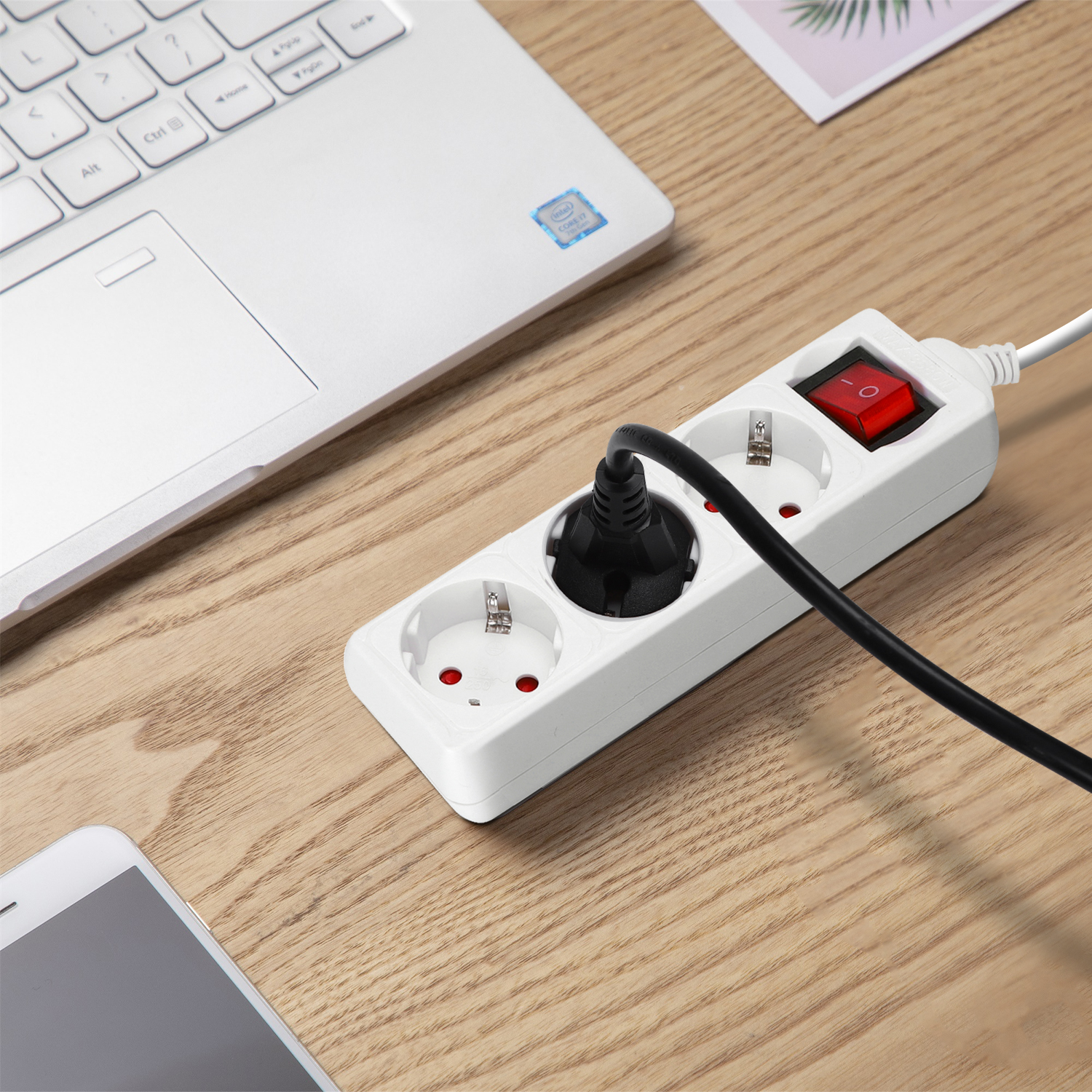 Weißes Verlängerungskabel mit rotem Schalter, schwarzem Stecker. Laptop und Telefon auf Holztisch.