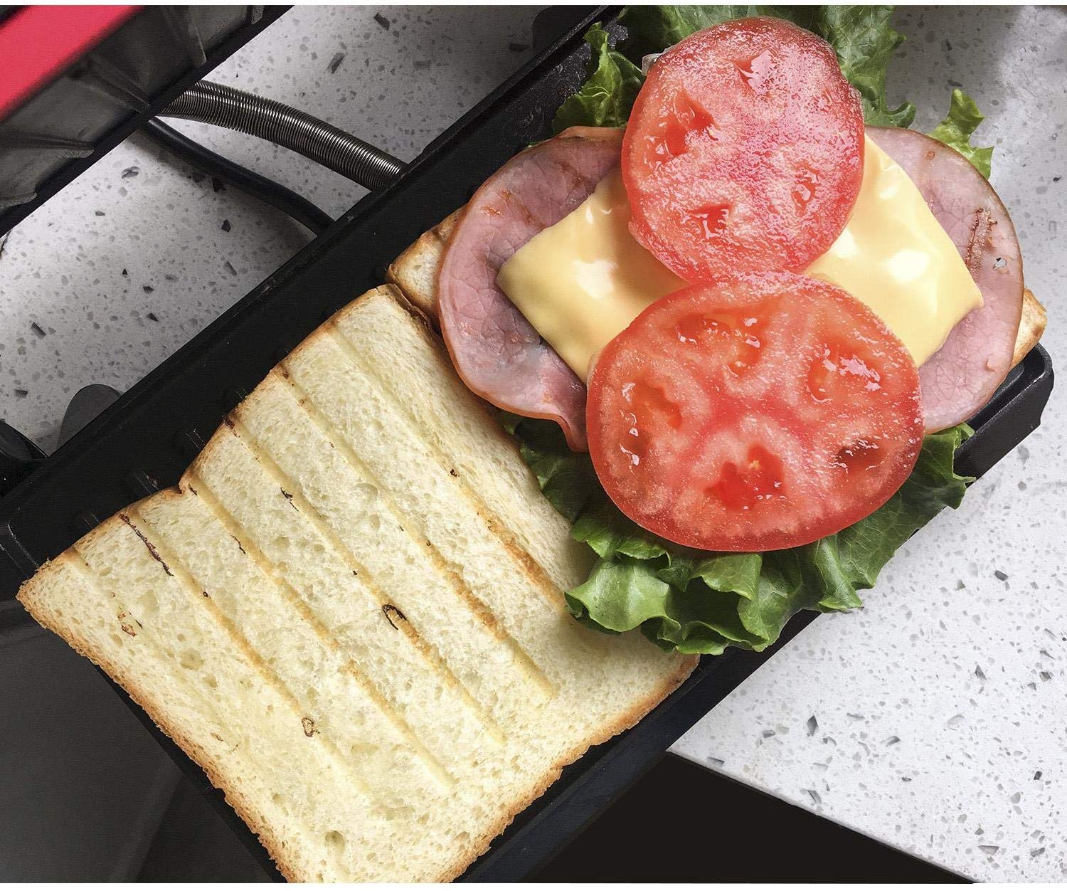 Panino su una griglia nera con pomodoro, formaggio, prosciutto e lattuga. Il pane è tostato.