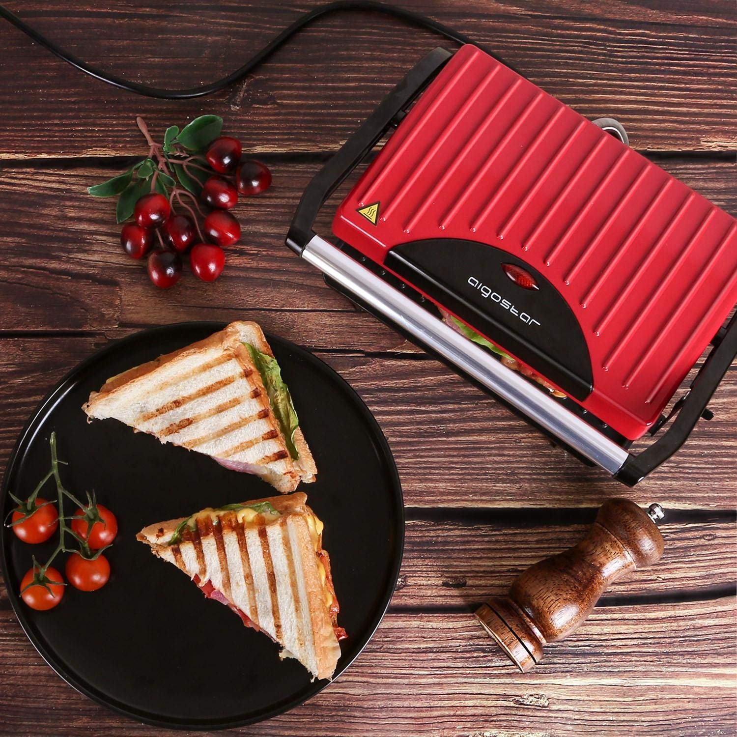 Piastra per panini rossa con cibo, pomodori e ciliegie su legno. Due panini e un macinapepe.