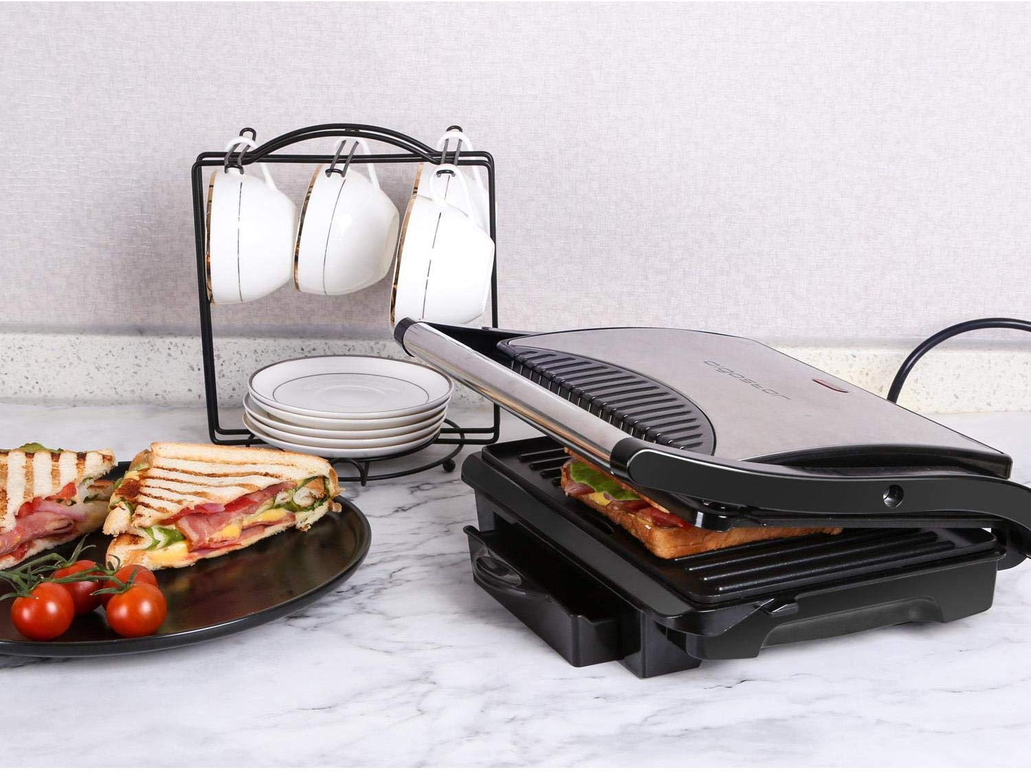 Macchina per sandwich con panini, pomodori e tazze su un tavolo. La macchina è aperta.