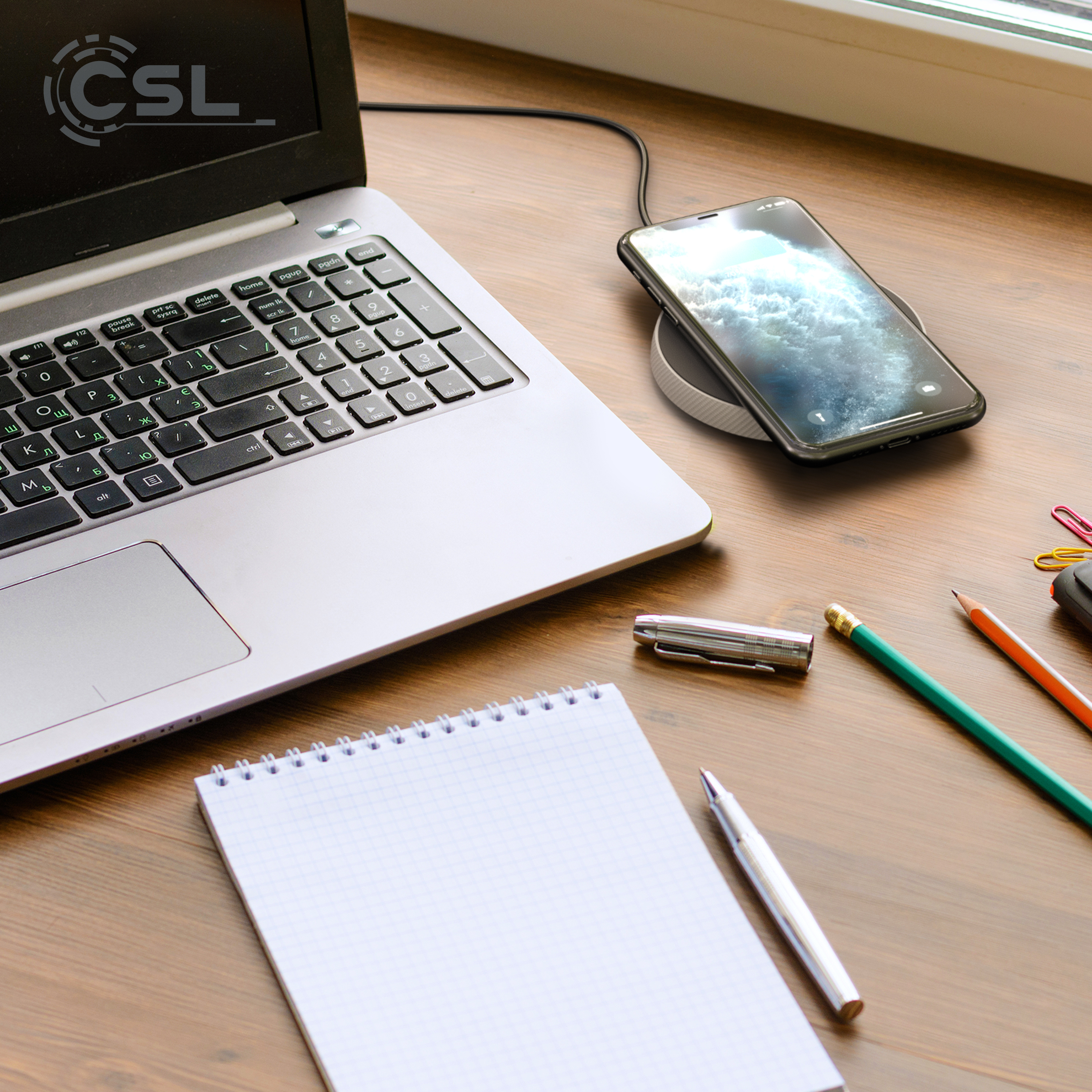 Laptop mit Telefon auf kabellosem Ladegerät. Notizblock, Stift, Bleistifte und Büroklammern auf dem Holzschreibtisch.
