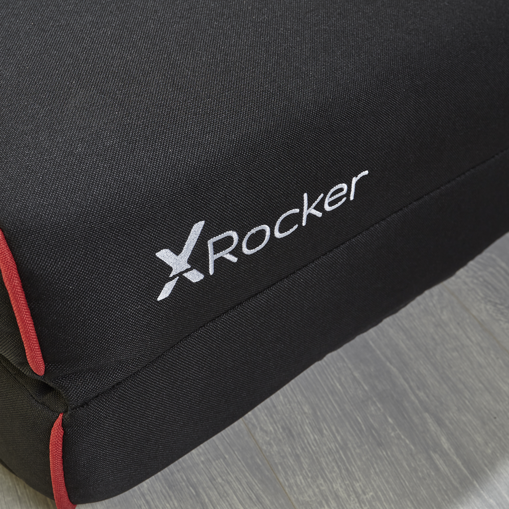 Schwarzes Stuhlkissen mit roter Umrandung und weißem "xRocker"-Logo. Das Kissen liegt auf einem Holzboden.