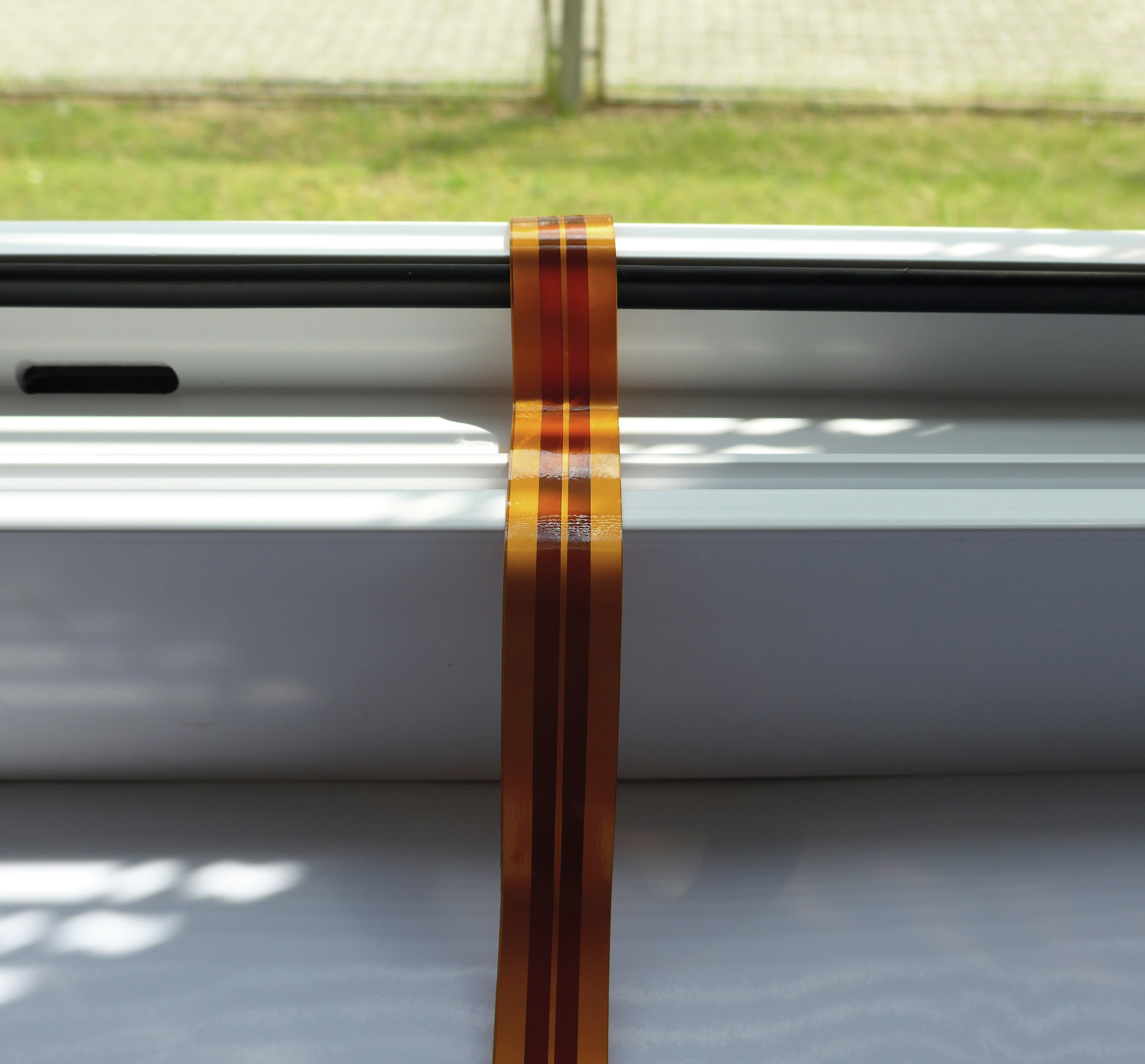 Ein flexibles Kabel mit kupferfarbenen Streifen liegt auf einem weißen Fensterrahmen.