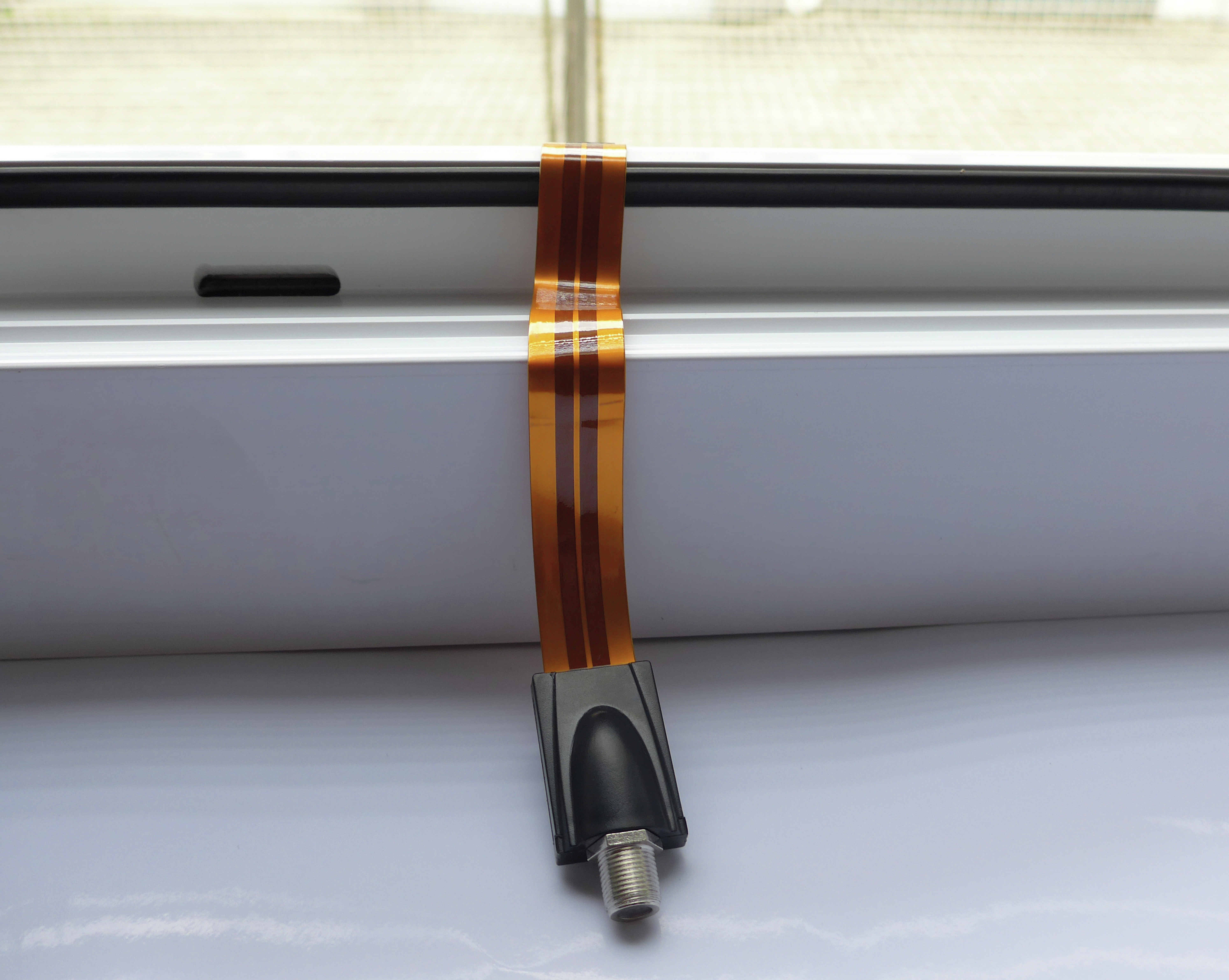 Flaches oranges Kabel mit schwarzem Stecker und silbernem Stecker, auf weißer Oberfläche am Fenster.