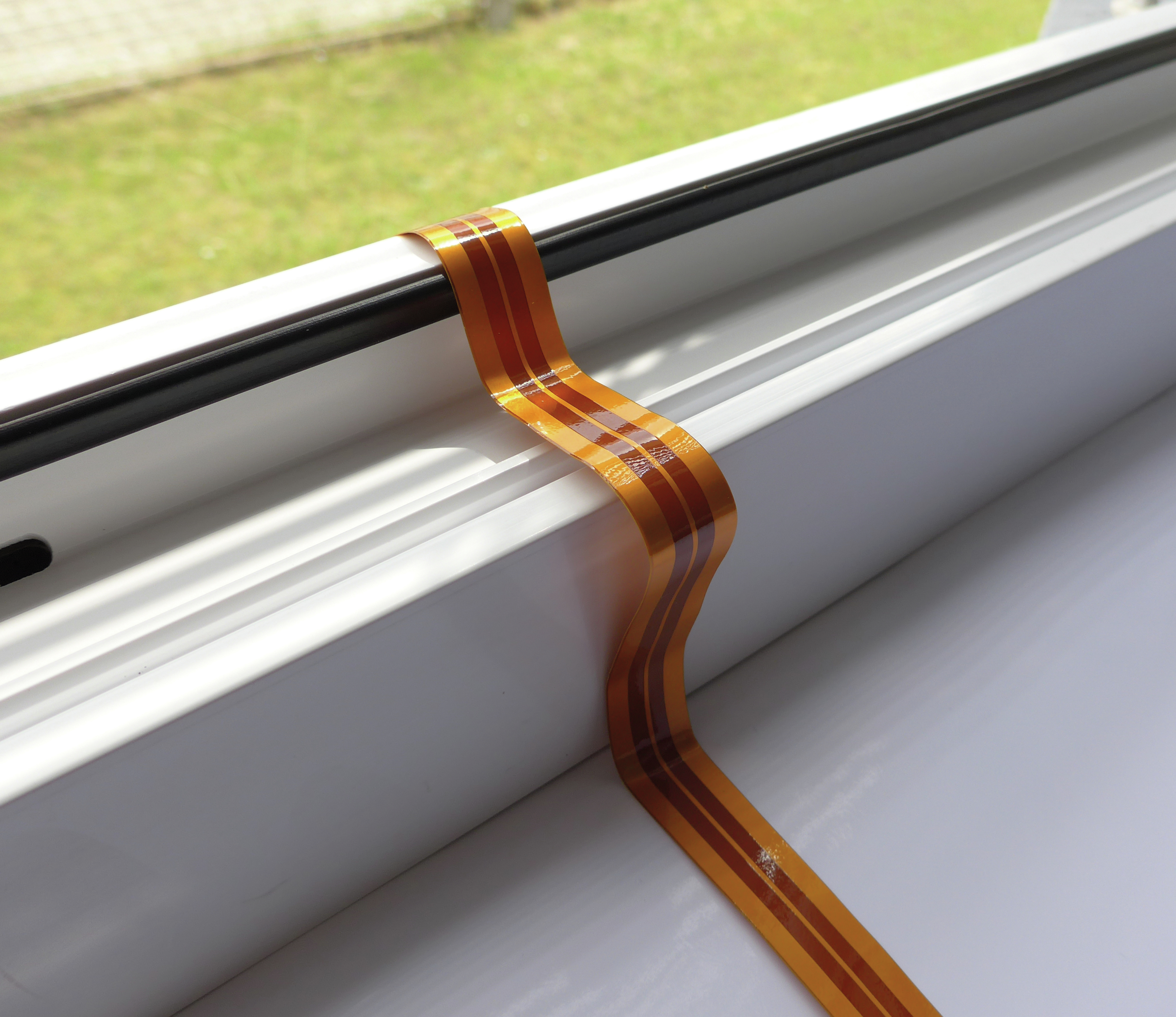 Ein flexibles, orangefarbenes Flachbandkabel, das über eine weiße Fensterbank verläuft, mit grünem Hintergrund.
