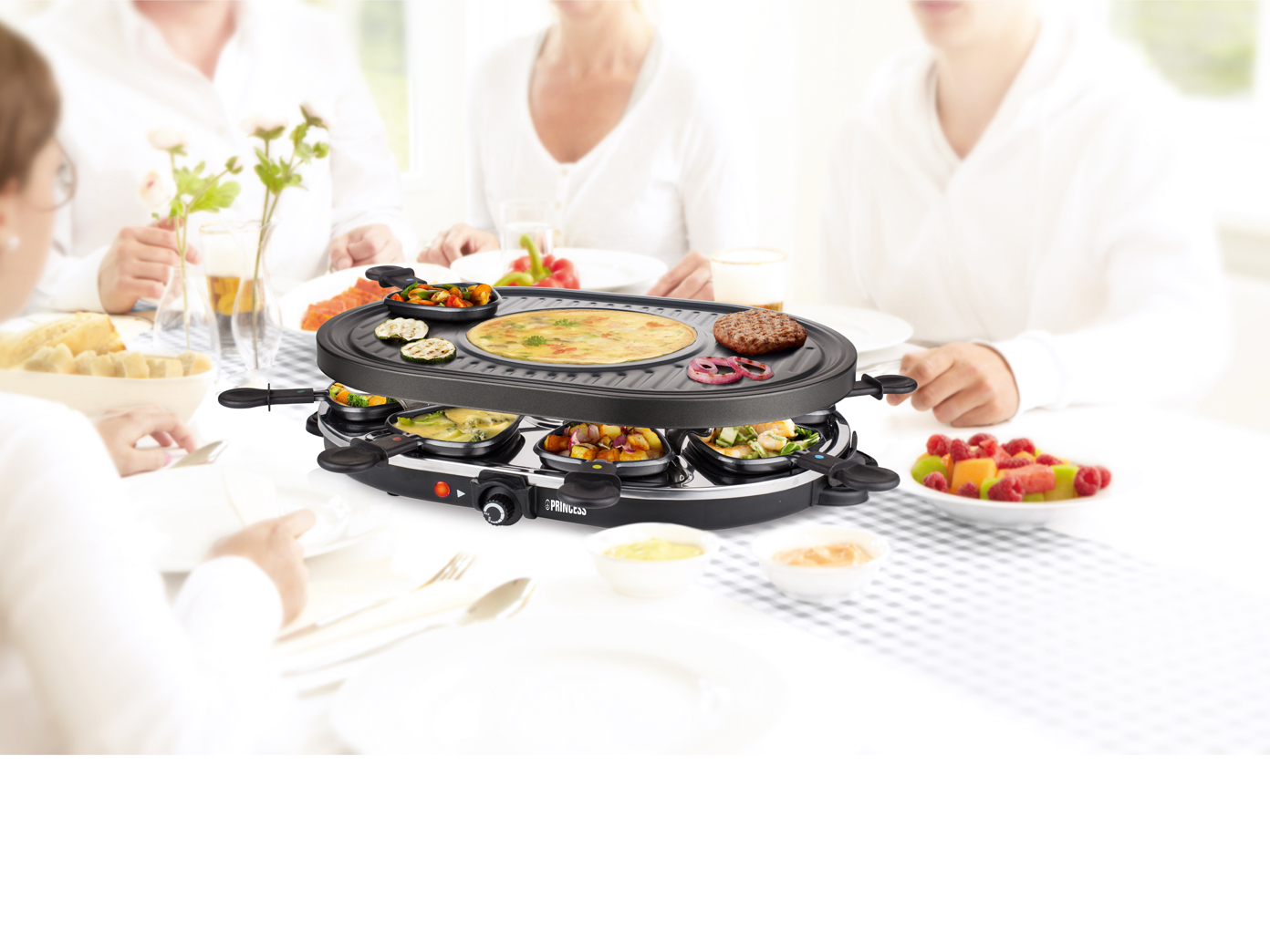 Raclette-Grill auf weißem Tisch mit Essen. Personen sitzen. Essen umfasst Gemüse und Fleisch.