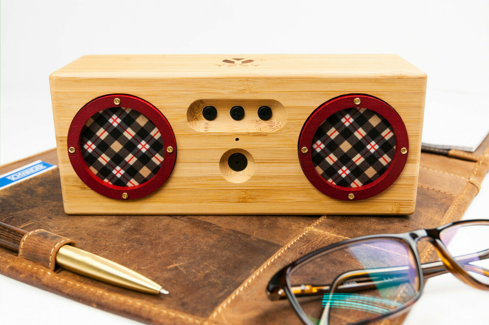 Holzlautsprecher mit roten Gittern. Er liegt auf einer Lederoberfläche, Stift und Gläser sichtbar.