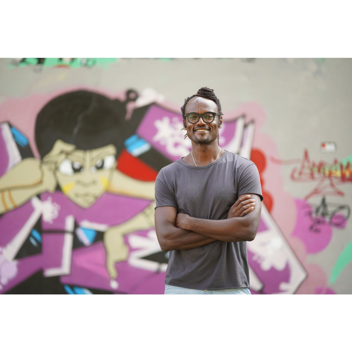 Ein Mann steht draußen mit verschränkten Armen vor einer Graffiti-Wand, trägt eine Brille und ein graues T-Shirt.