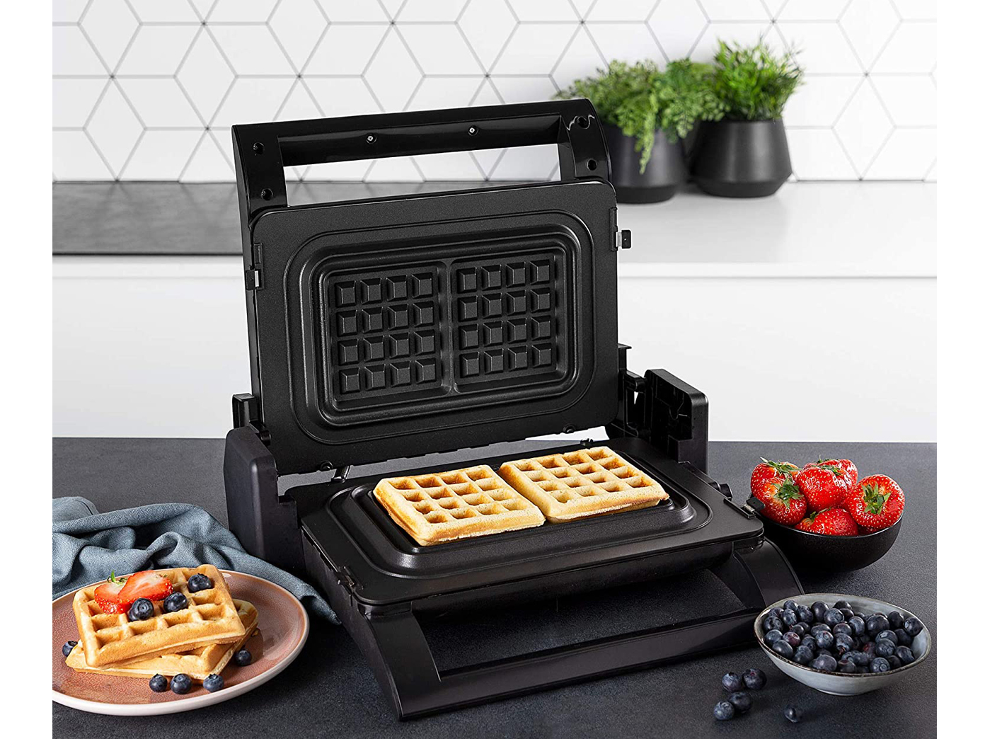Waffeleisen mit Waffeln drin und Waffeln mit Beeren auf einem Teller und Blaubeeren.