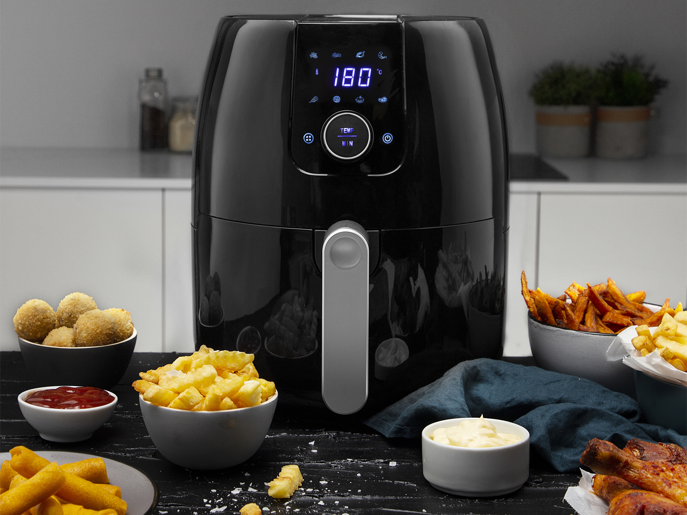 Schwarze Heißluftfritteuse mit Essen darum. Pommes, Nuggets und Saucen sind sichtbar.