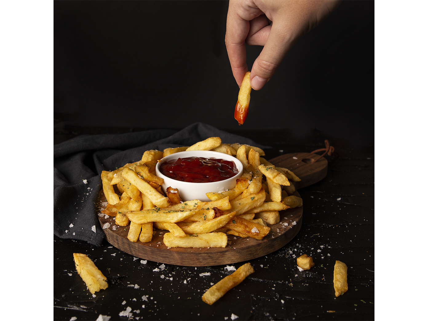 Ręka maczająca frytkę w misce z ketchupem. Frytki na drewnianej desce. Ciemne tło.