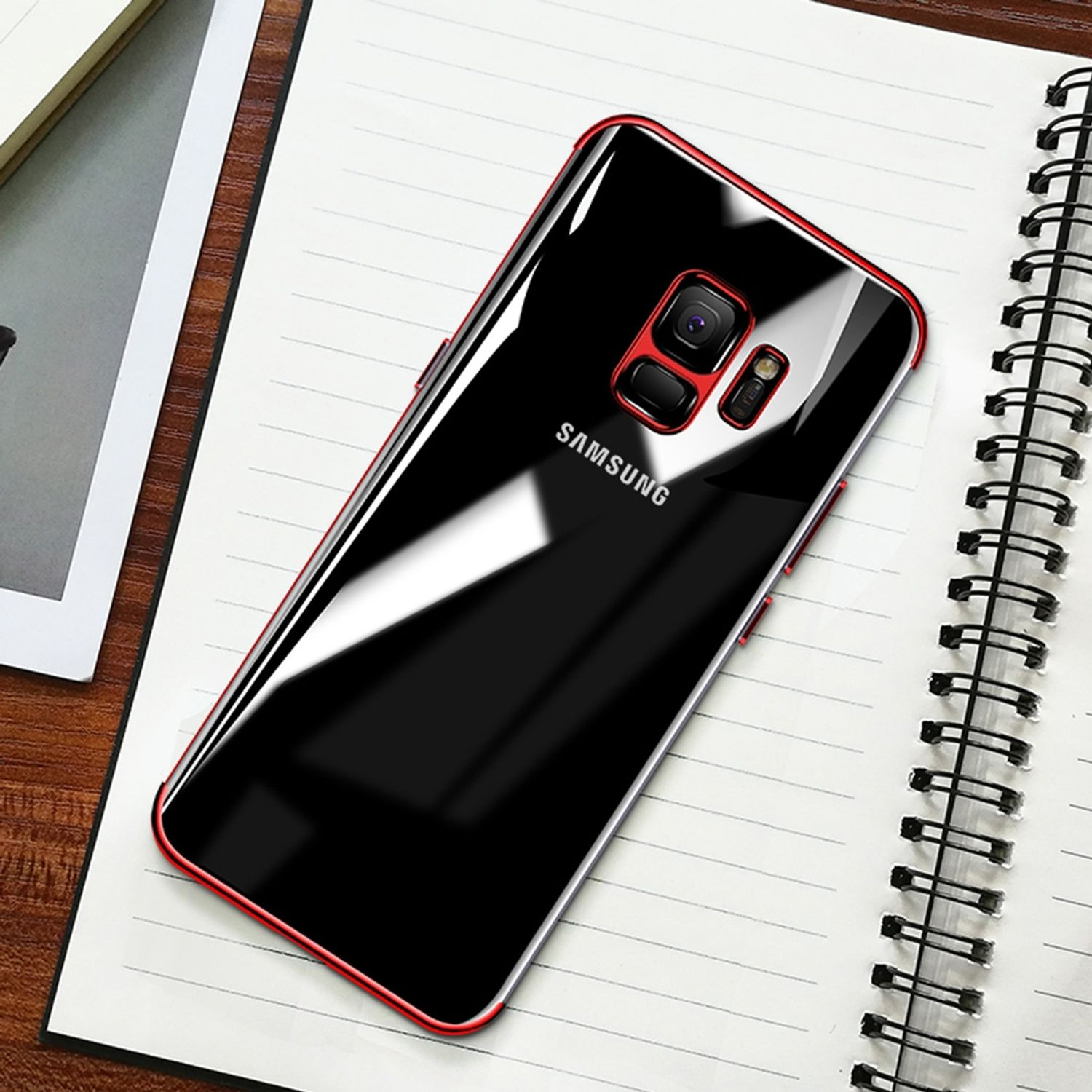Ein schwarzes Telefon in einer rot umrandeten Hülle auf einem Notizbuch. Hülle hat Kameraausschnitt und Samsung Logo.