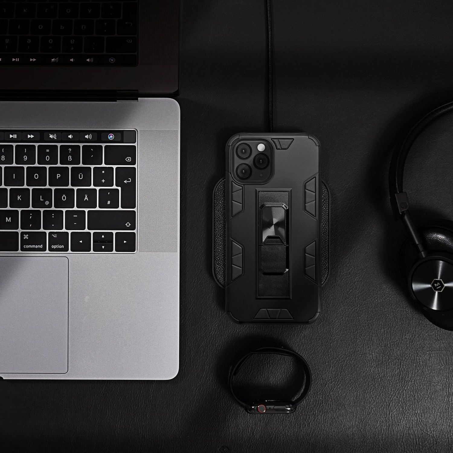 Schwarze Handyhülle mit Ständer, Apple Watch, Kopfhörer und Laptop auf schwarzer Oberfläche.