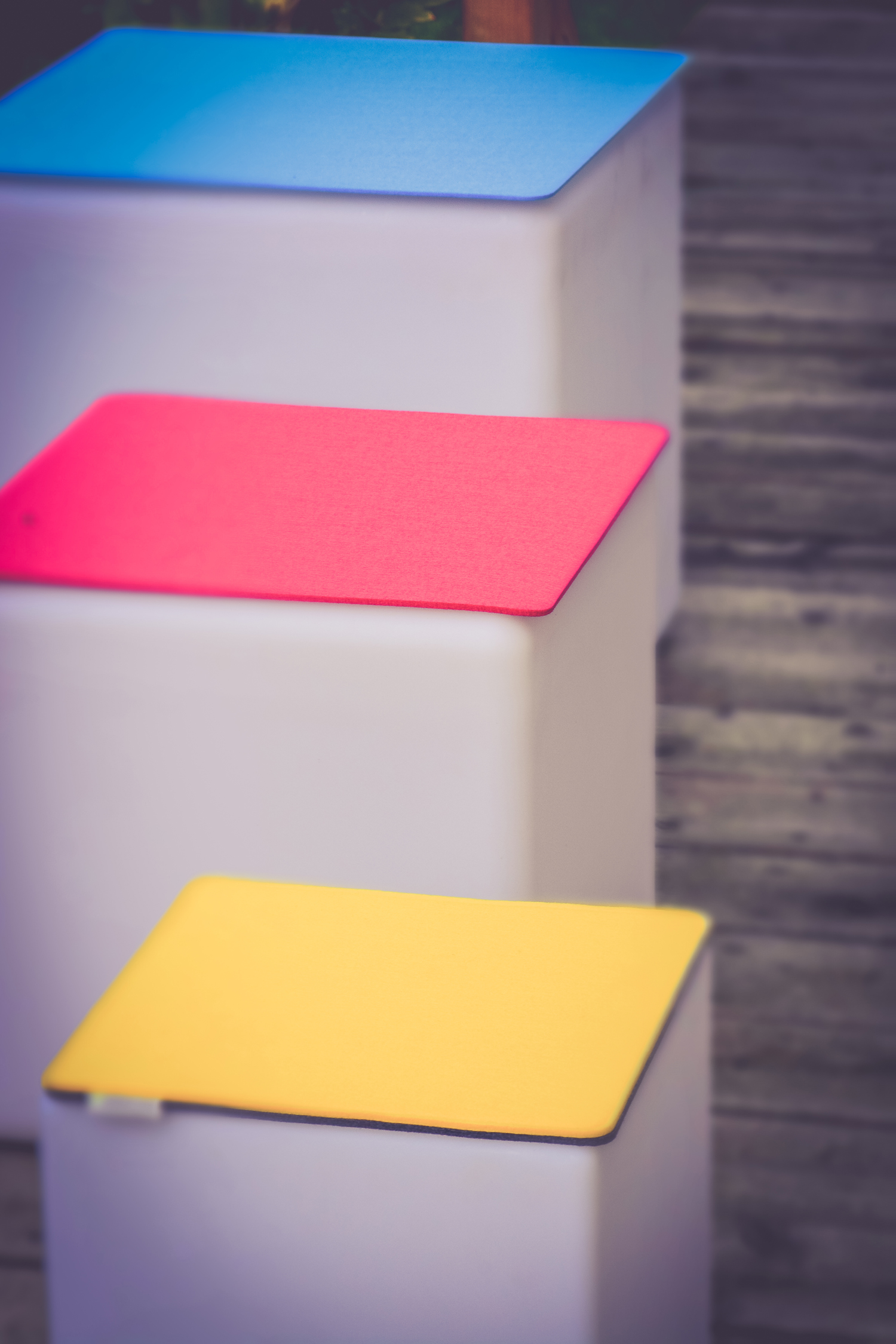 Drei weiße Boxen mit farbigen Deckeln: blau, rot und gelb. Sie sind draußen auf einer hellen Oberfläche.