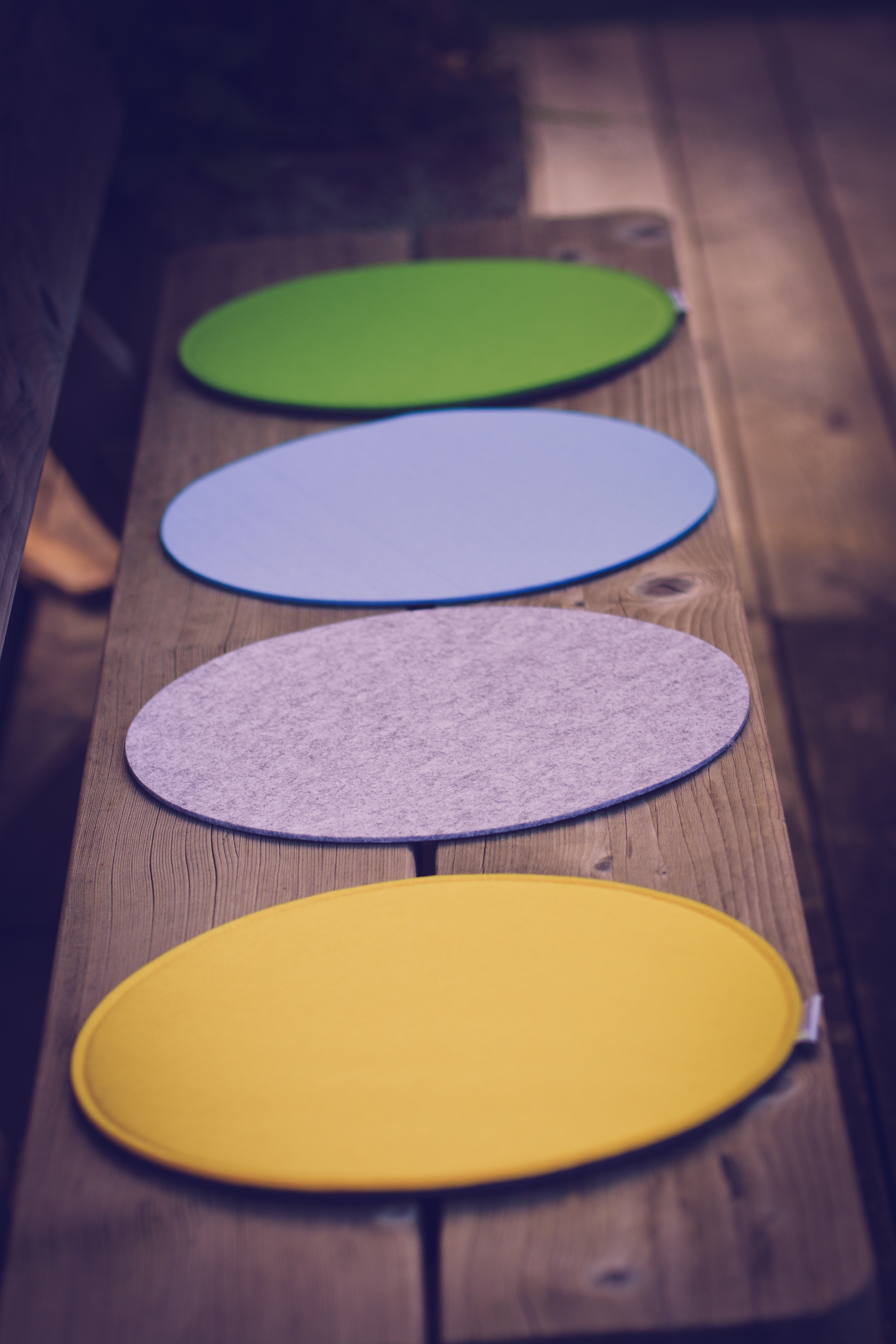 Vier ovale Filz-Tischsets in Grün, Blau, Grau und Gelb auf einer Holzbank.