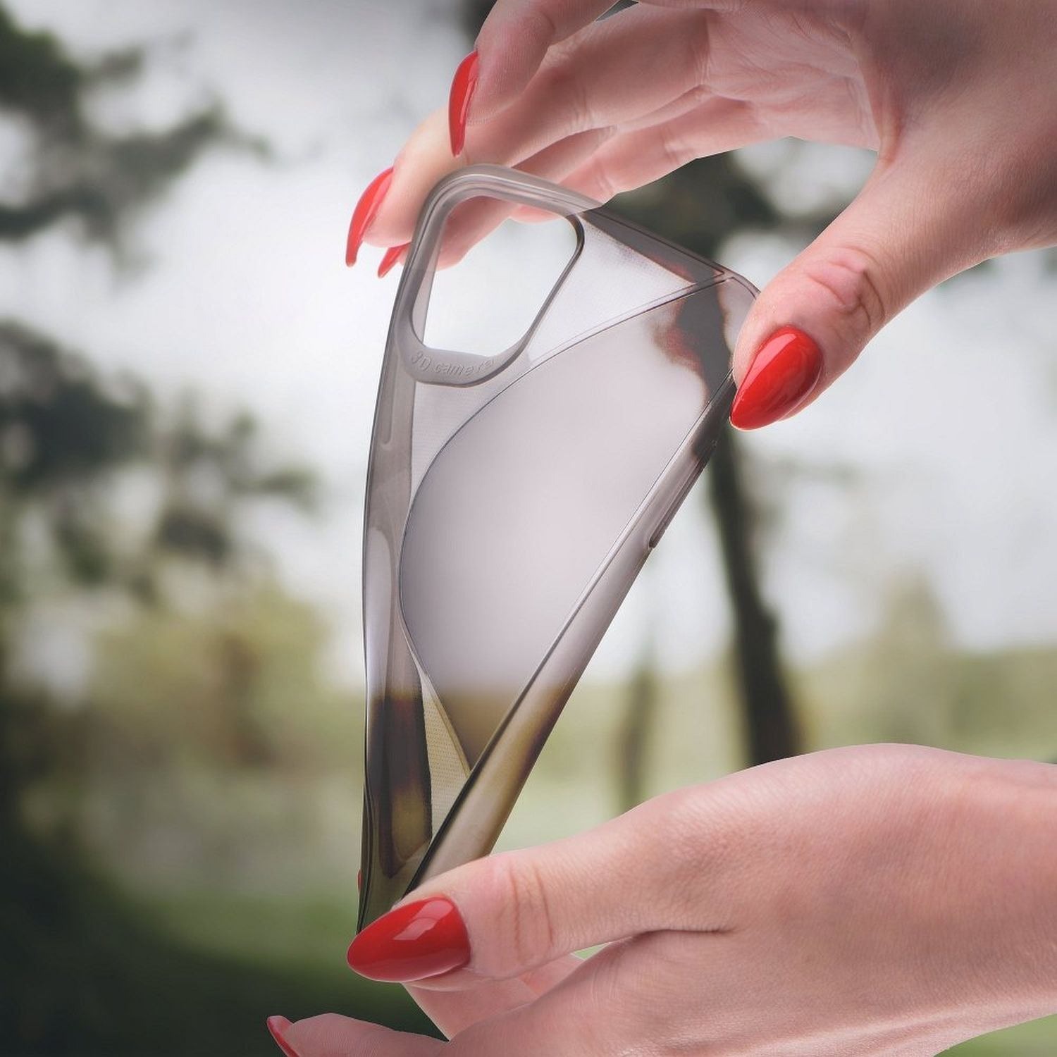 Hände halten eine transparente Handyhülle, Flexibilität zeigend. Roter Nagellack. Verschwommener grüner Hintergrund.