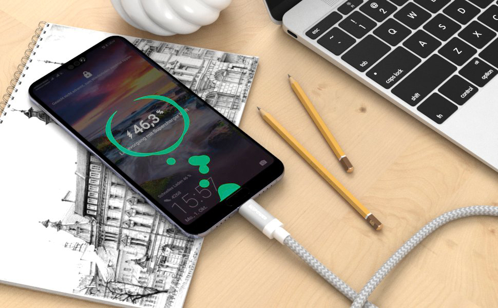 Telefon wird mit Kabel geladen. Bleistifte und ein Notizbuch liegen auf einer Holzoberfläche.