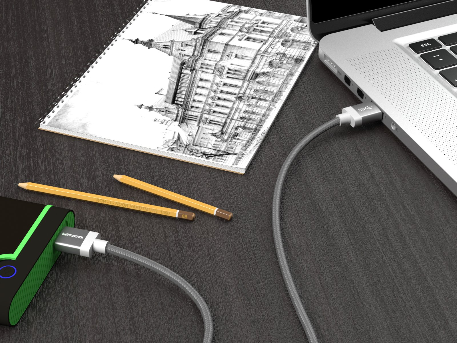 Ein Laptop ist über ein Kabel mit einer Powerbank verbunden. Ein Notizbuch und Bleistifte sind vorhanden.