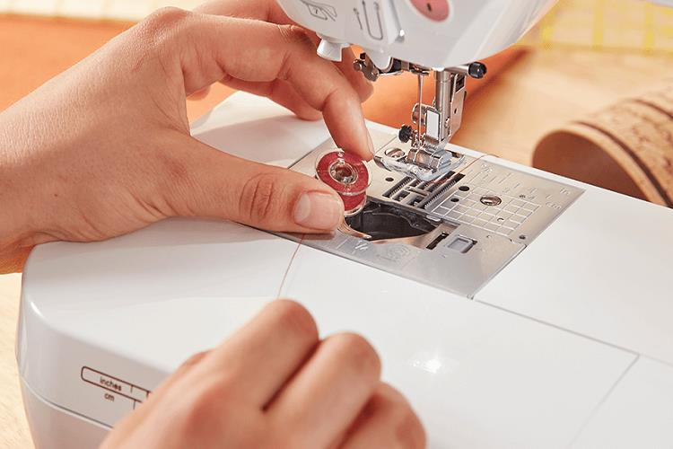 Hände legen eine rote Spule in eine weiße Nähmaschine. Die Nähmaschine ist an.