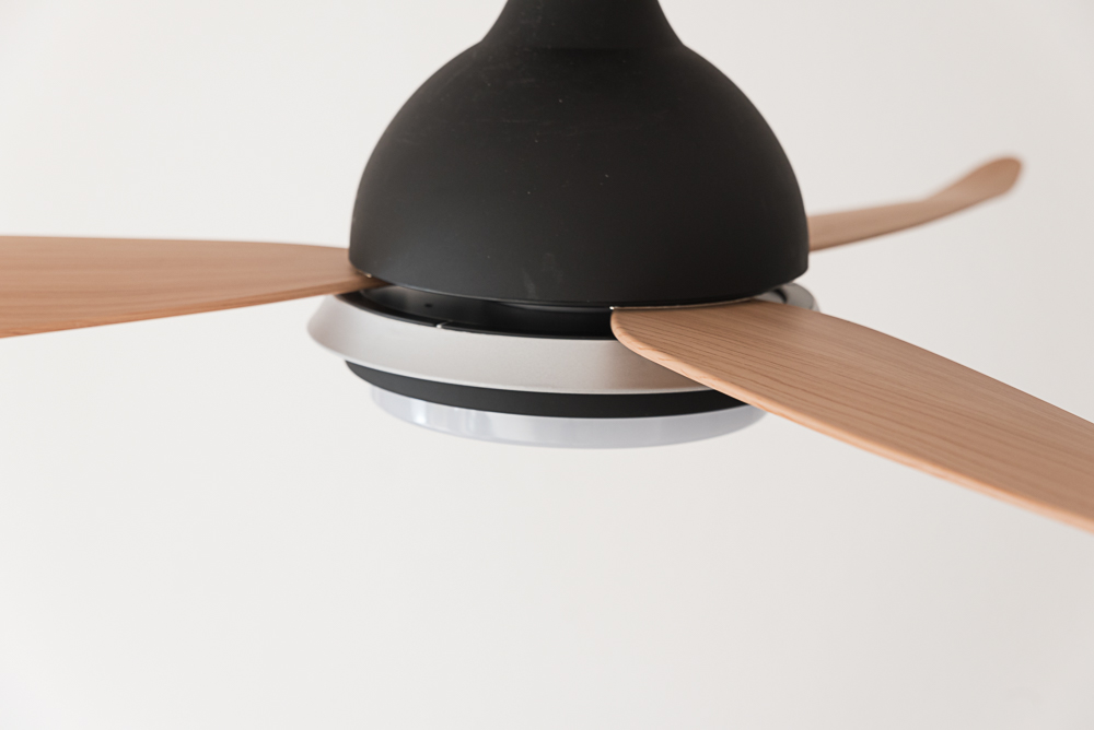 Deckenventilator mit drei Holzflügeln und einem schwarzen Gehäuse mit silberner Zierleiste. Weißer Hintergrund.