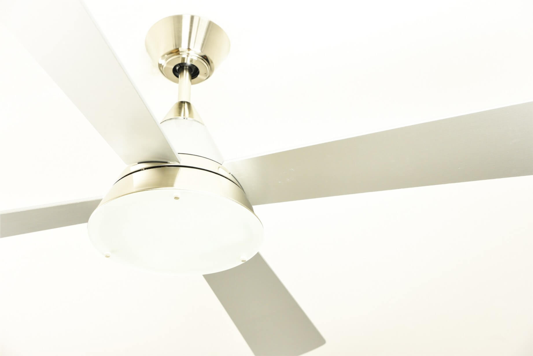 Un ventilatore da soffitto con quattro pale argentate e una base bianca, contro un soffitto bianco.