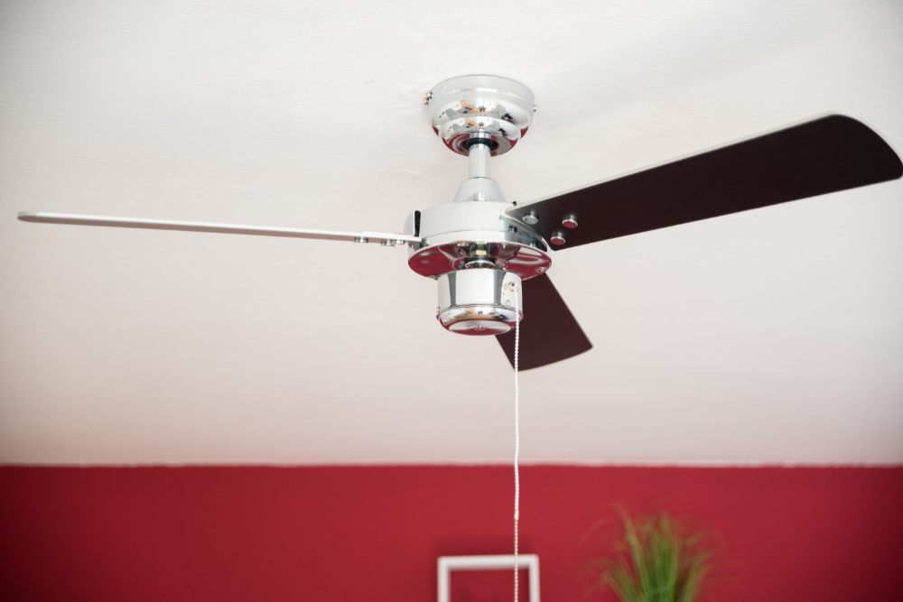 Ein Deckenventilator mit schwarzen Flügeln und einem Chrommotorgehäuse, vor rotem und weißem Hintergrund.