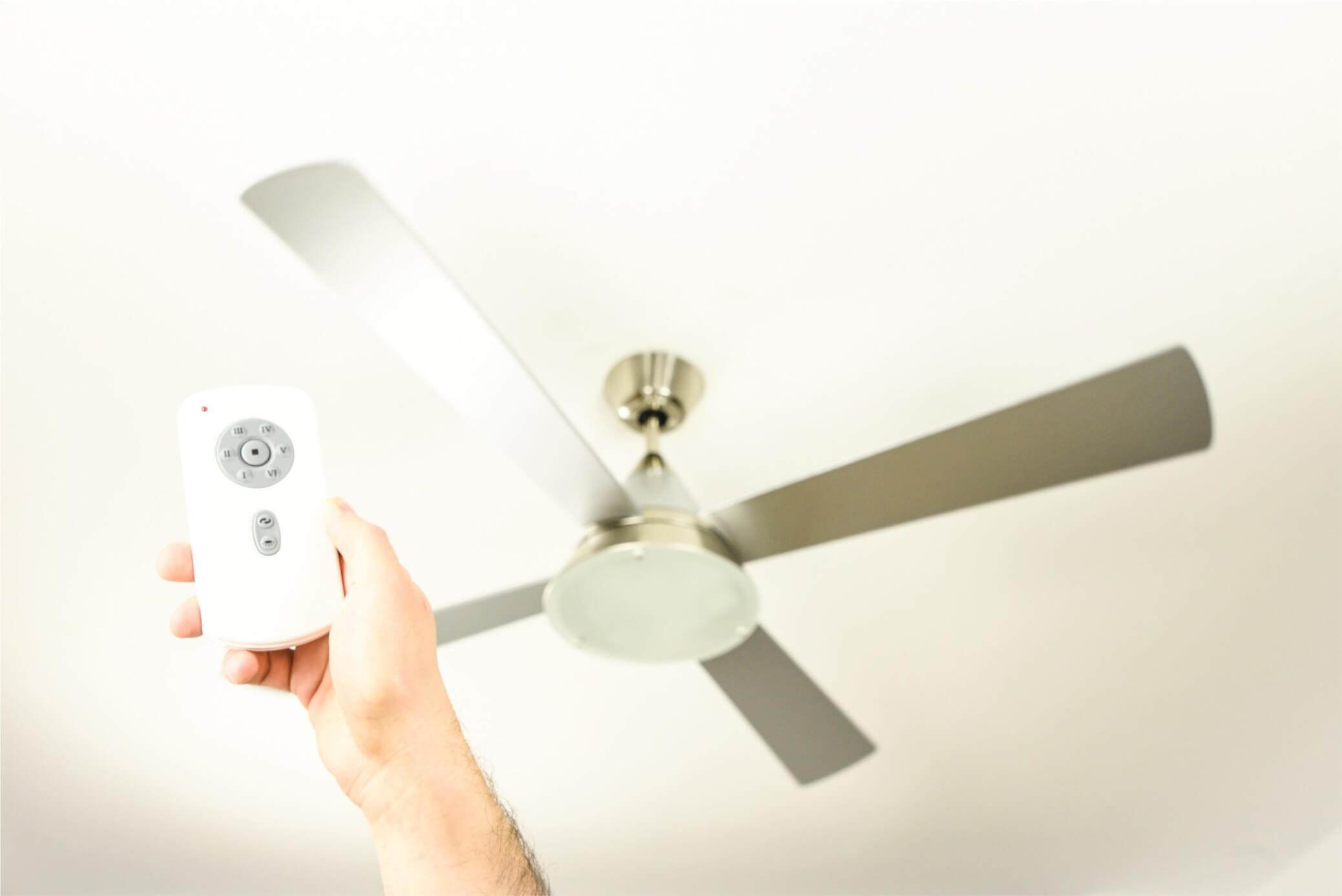 Una mano che tiene un telecomando bianco verso un ventilatore da soffitto con pale argentate.