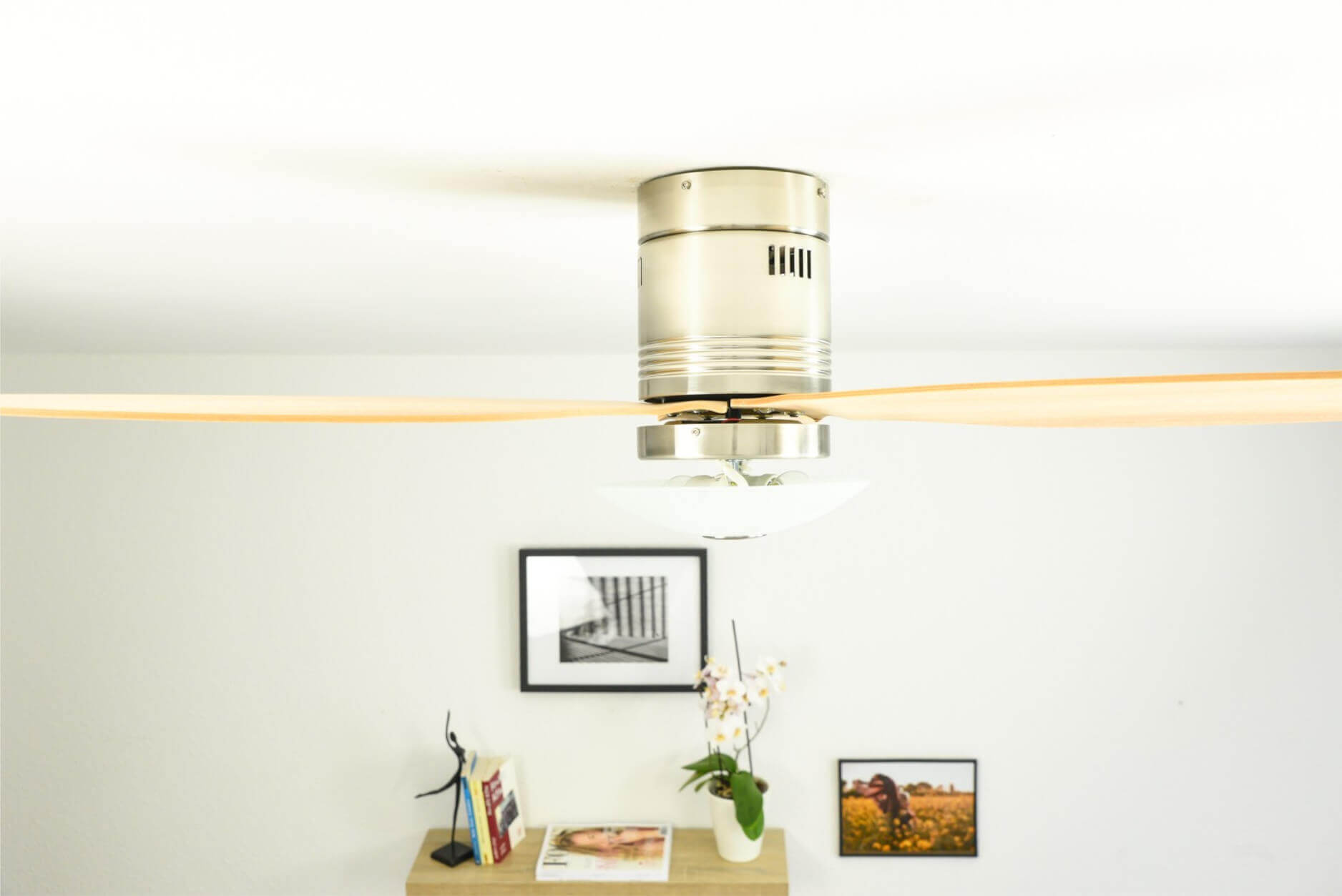 Deckenventilator mit Holzblättern, unter weißer Decke, mit Bilderrahmen und Dekor.