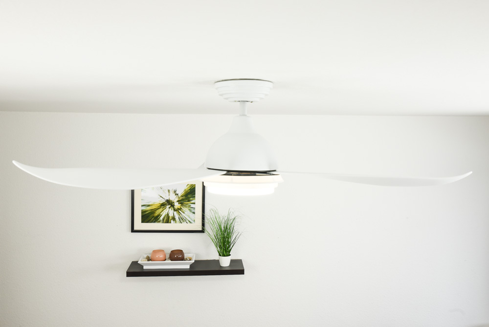 Weißer Deckenventilator mit gebogenen Blättern, einer Leuchte und Dekoration an der Wand darunter.