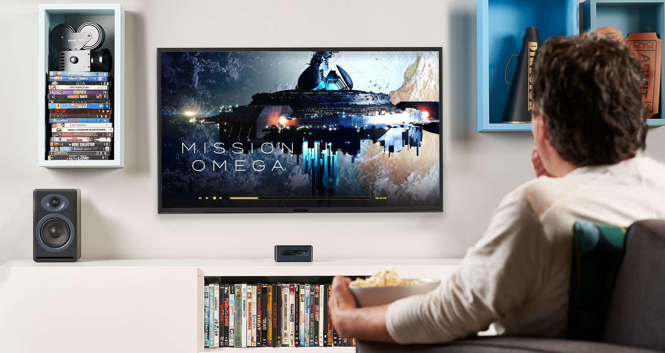 Ein Mann sieht fern. Der Bildschirm zeigt einen Film mit dem Titel Mission Omega. Es gibt Regale und Lautsprecher.