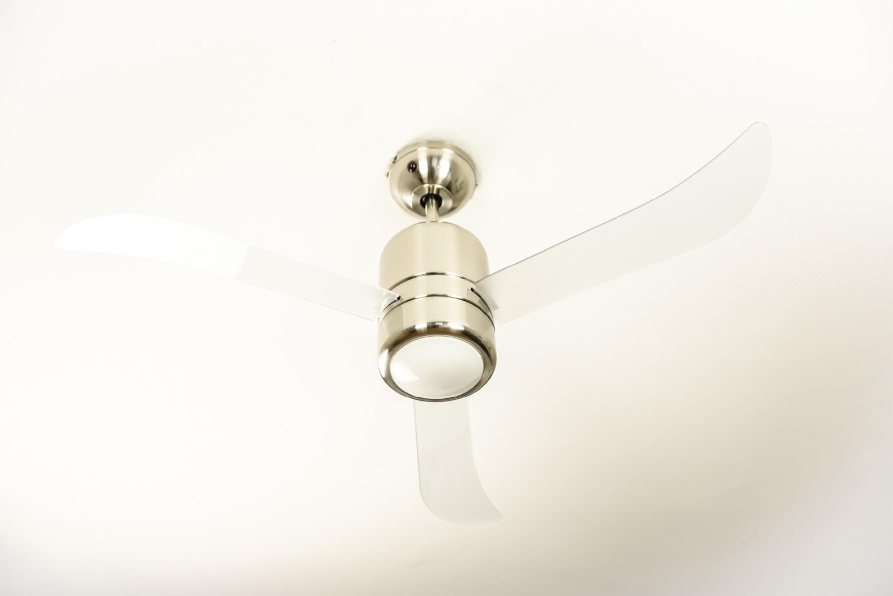Deckenventilator mit drei transparenten Flügeln und einem Licht. Es ist silberfarben.