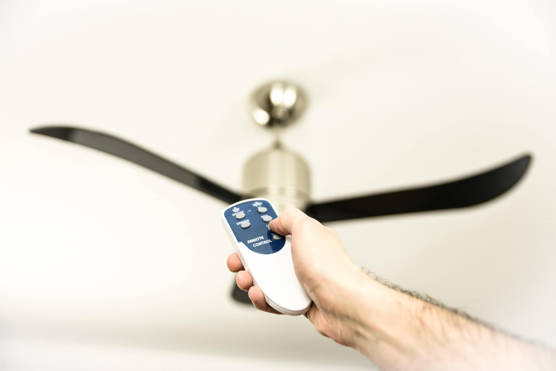 Hand hält Fernbedienung, gerichtet auf einen Deckenventilator mit schwarzen Flügeln.