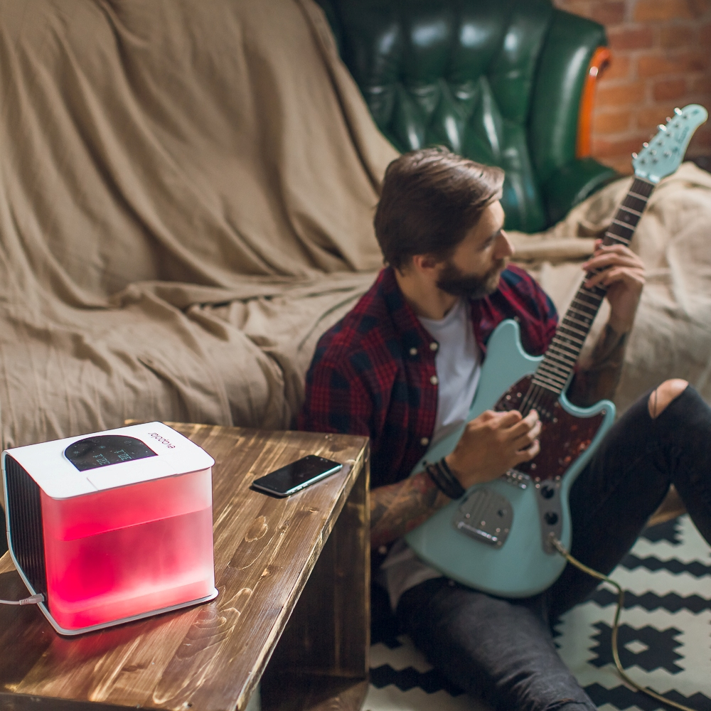 Mann spielt blaue Gitarre nahe Licht. Das Licht ist rot. Telefon auf Holztisch. Couch im Hintergrund.