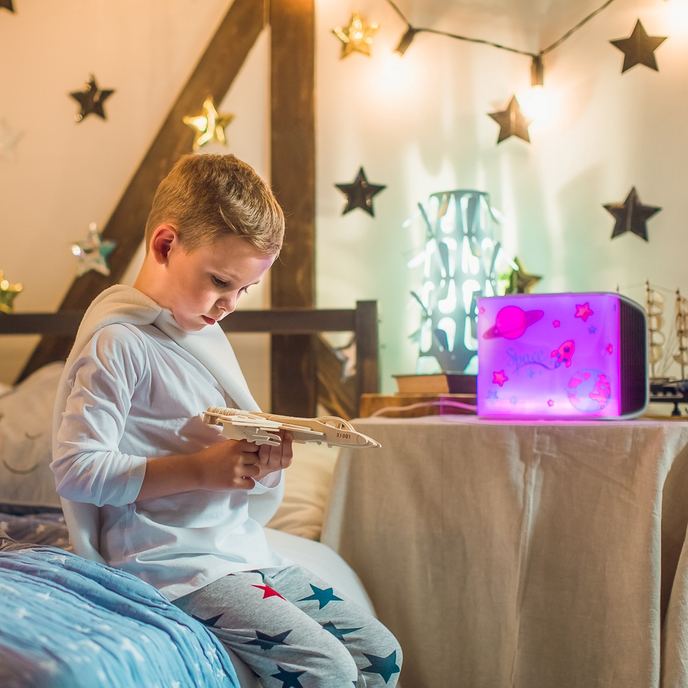 Ein Junge sitzt auf einem Bett mit einem Spielzeugflugzeug, einer Lampe und Sternen. Der Raum ist beleuchtet.
