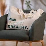 Eine Tragetasche aus Canvas mit den Worten "TAKE IT BREATHY" darauf, die auf einem grünen Stuhl liegt.
