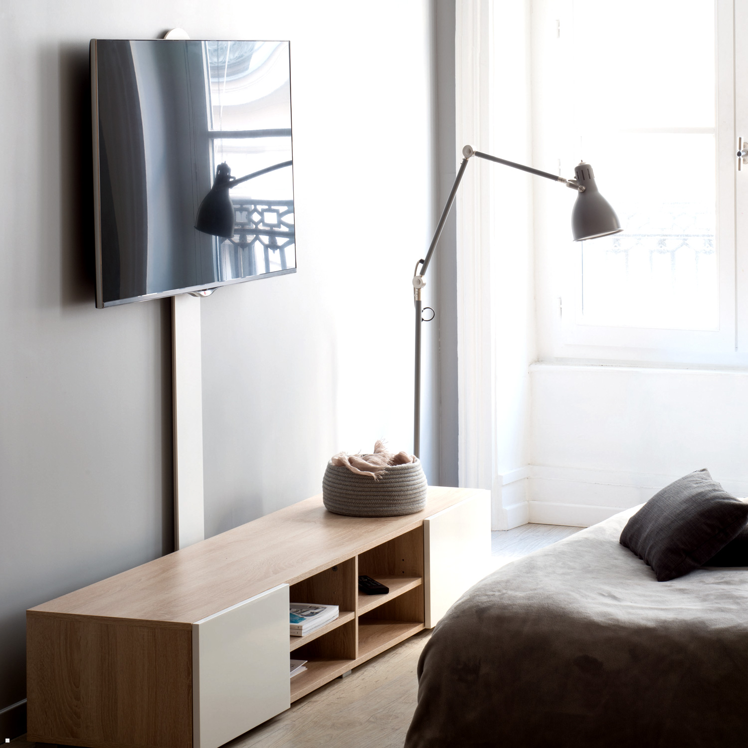 Una scena di camera da letto con un muro grigio, una TV, una lampada e un letto con cuscino e coperta.
