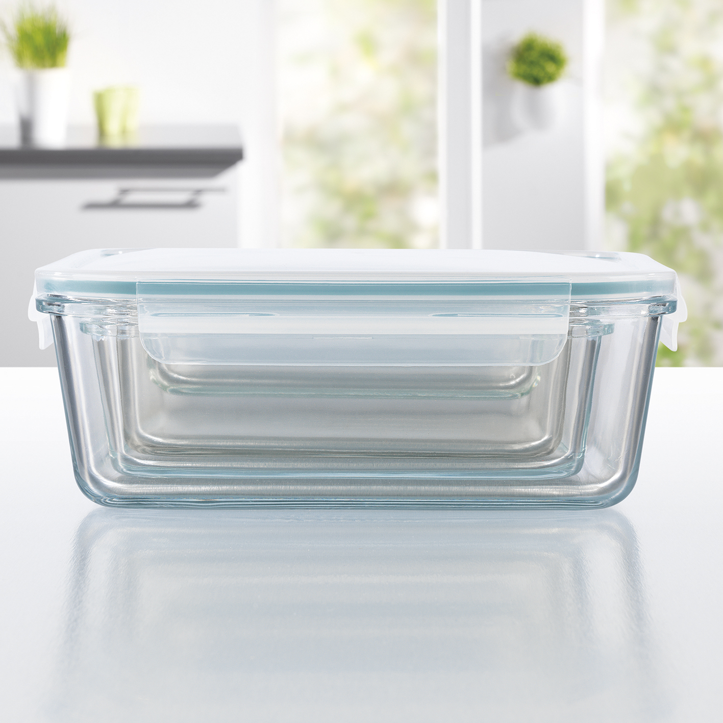 Drei transparente Glas-Aufbewahrungsbehälter mit Deckeln, gestapelt auf einer weißen Oberfläche, mit Küchenhintergrund.