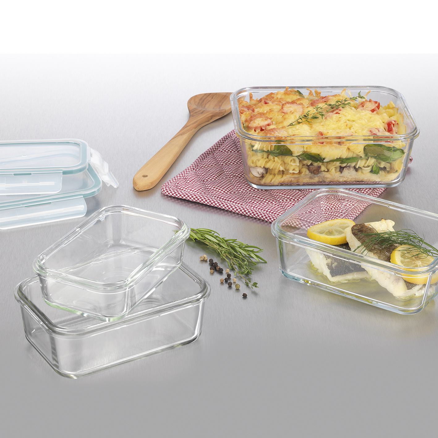 Glas-Lebensmittelbehälter mit gekochtem Essen, ein Holzloeffel und Kräuter auf silberner Oberfläche.