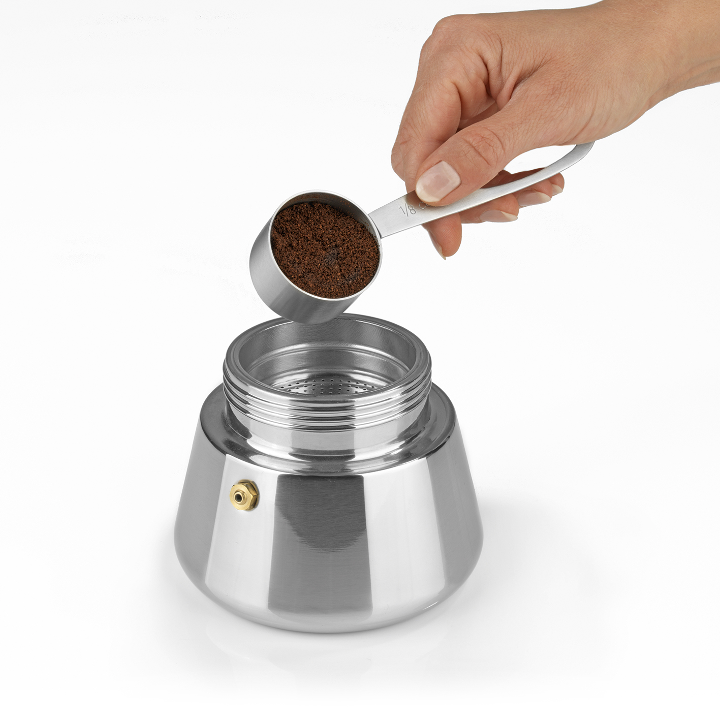 Eine Person schüttet Kaffeepulver aus einem Löffel in eine Espressokanne aus Edelstahl. Weißer Hintergrund.