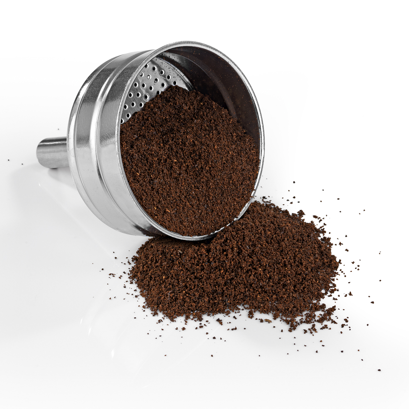 Ein Kaffeefilter aus Chrom, bei dem Kaffeepulver auf eine weiße Oberfläche verschüttet wird.