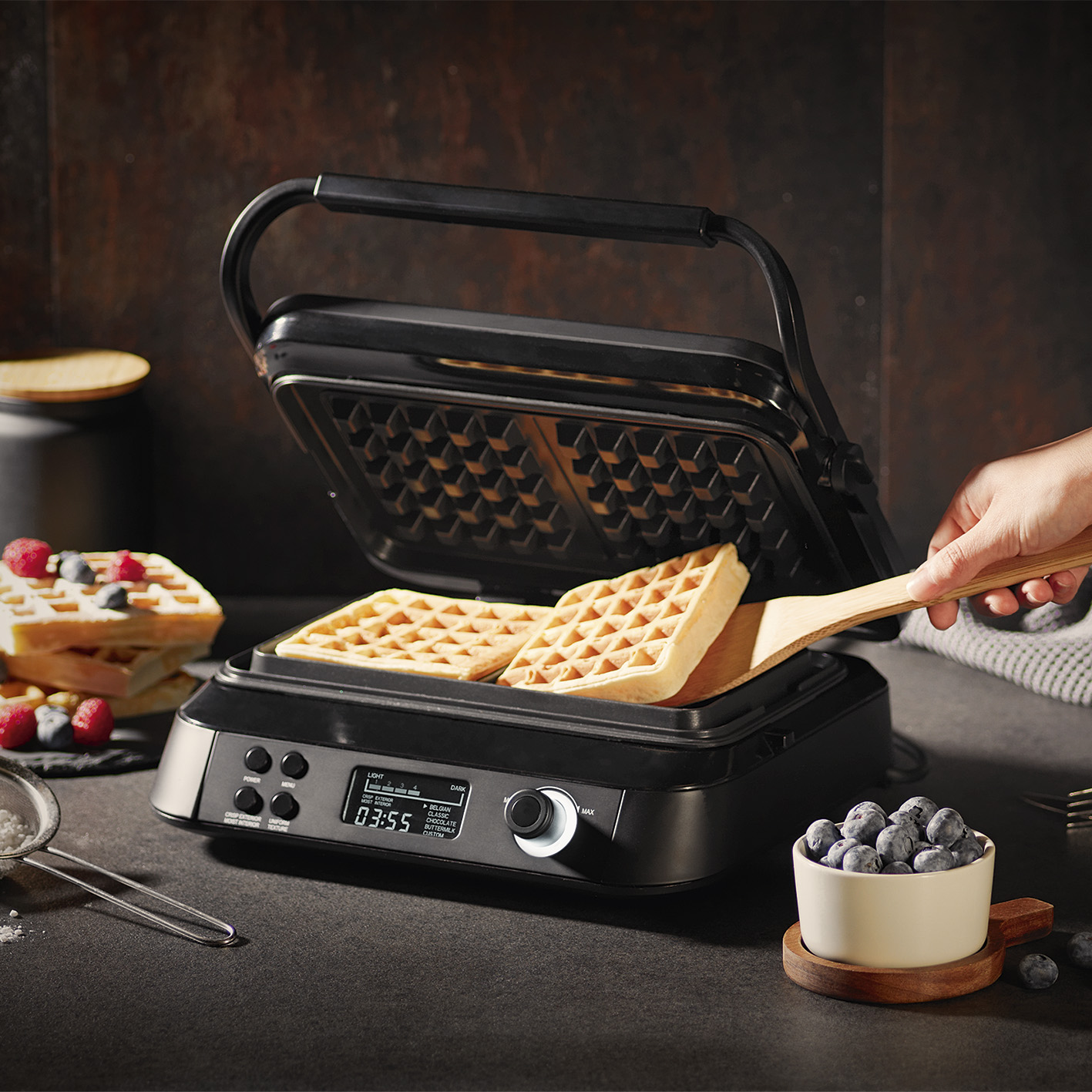 I waffle sono in una macchina per waffle, con una spatola di legno. Mirtilli e waffle sono sullo sfondo.