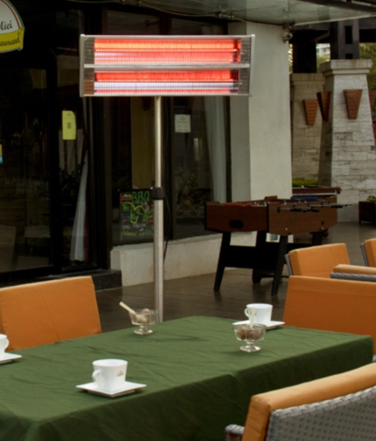Restaurant im Freien. Tische mit grünen Tischdecken, Stühlen und Heizung. Im Hintergrund ein Tischfußball.