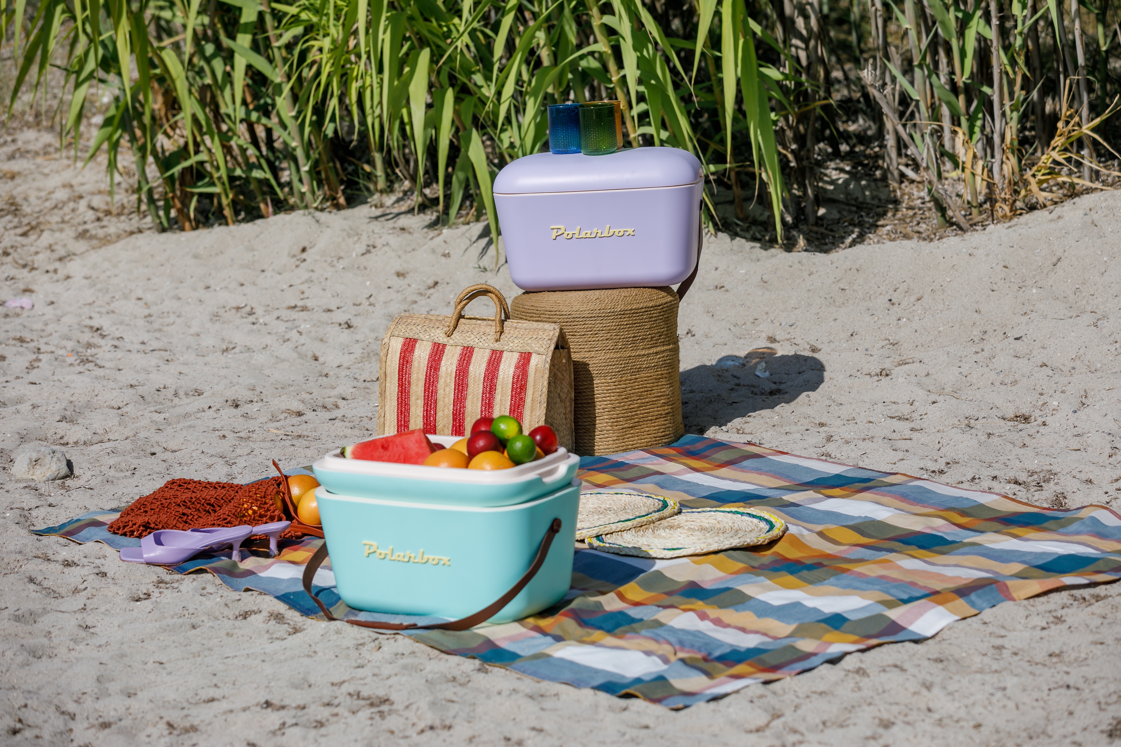 Scena picnic sulla sabbia: scatole, cesto, cibo, coperta, sfondo spiaggia.