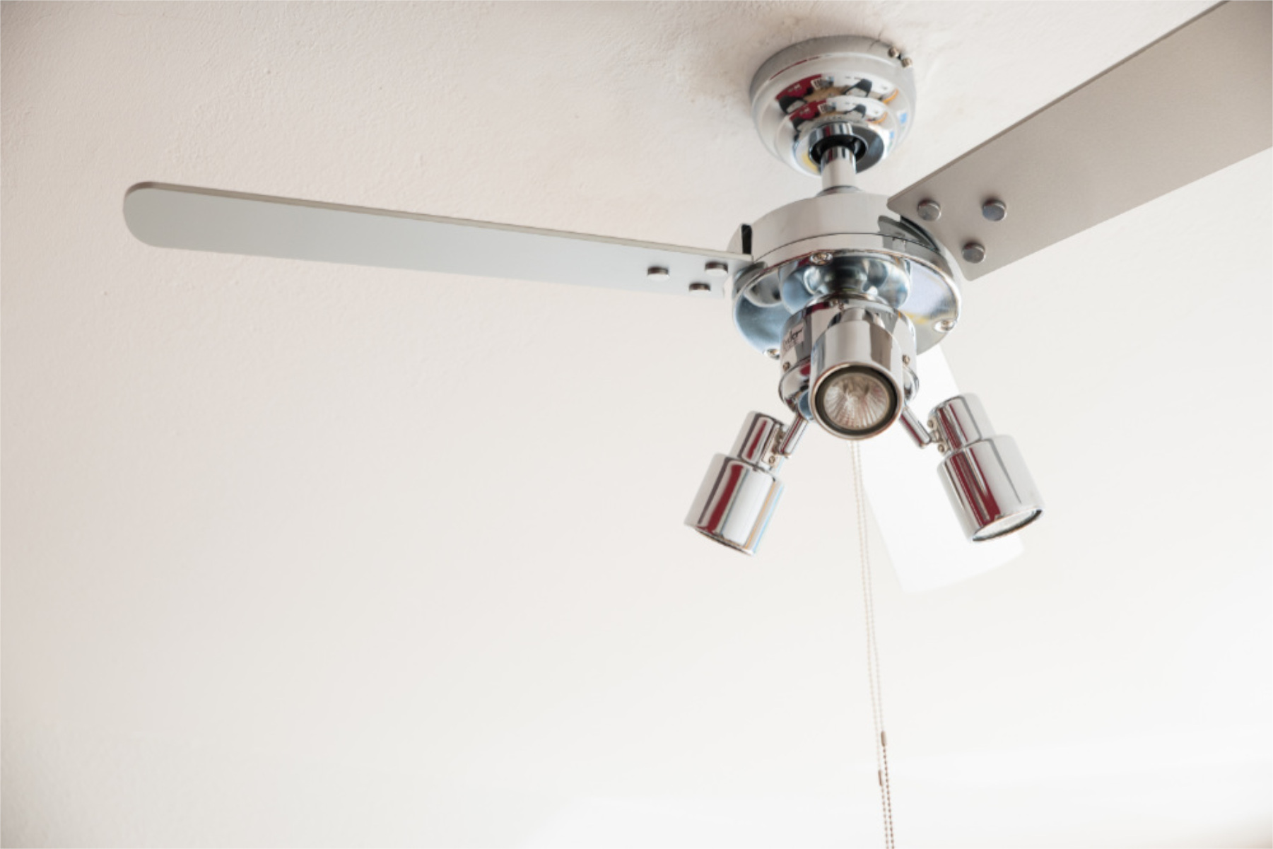 Deckenventilator mit drei Blättern und drei Strahlern. Der Lüfter ist verchromt, die Decke weiß.