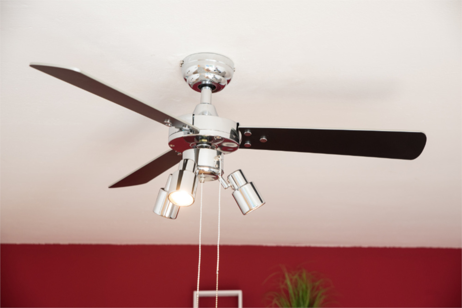 Ein Deckenventilator aus Chrom mit drei Lichtern darunter. Der Hintergrund ist eine rote Wand und Decke.