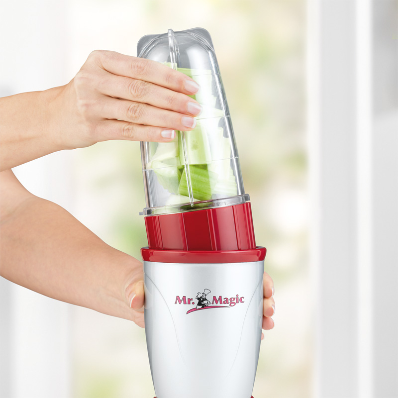 Mixer mit roten Teilen. Eine Hand legt grünes Gemüse hinein.