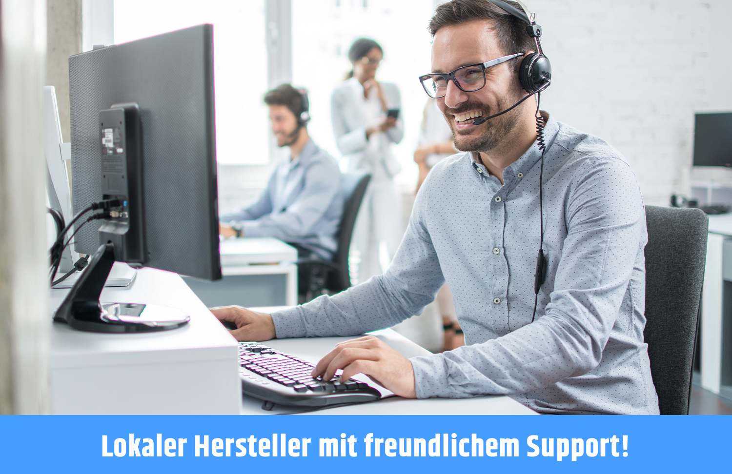 Ein Callcenter-Mitarbeiter mit Headset, lächelnd und am Computer arbeitend, mit Kollegen im Hintergrund.