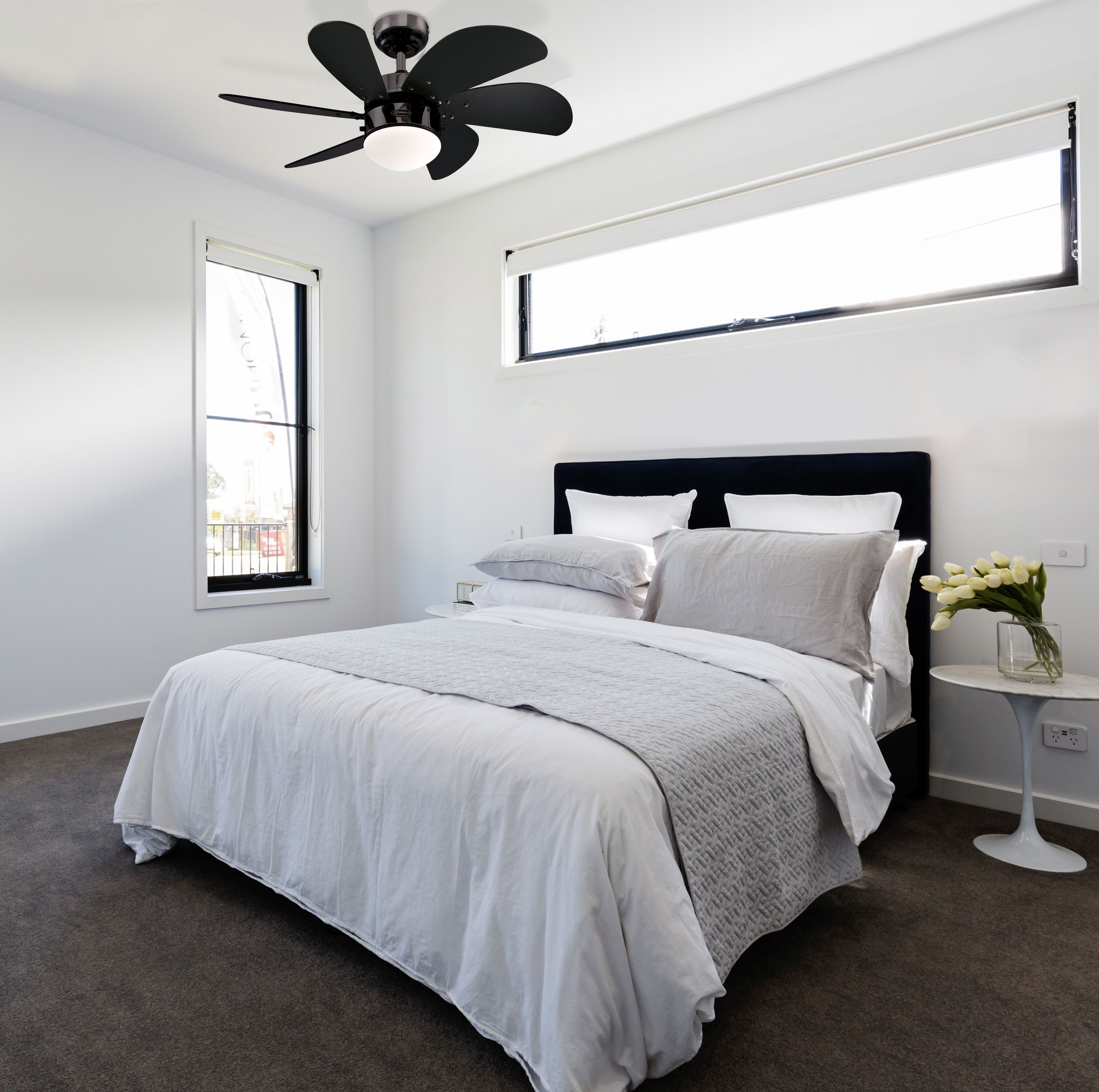Schlafzimmer mit weißen Wänden, Bett mit grauer Decke, weißen Kissen und schwarzem Deckenventilator. Fenster im Hintergrund.