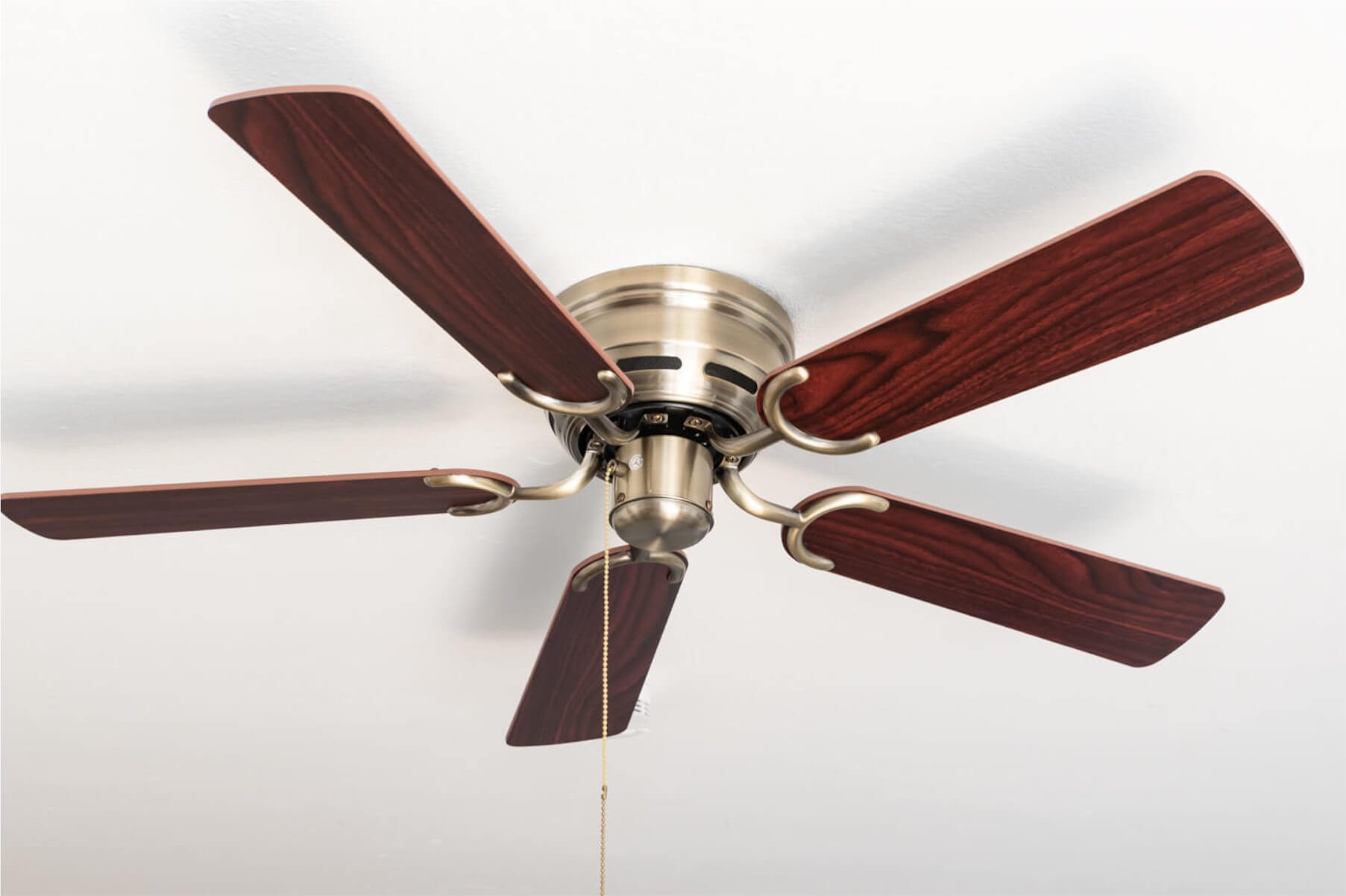 Deckenventilator mit 5 Holzblattflügeln, Messingakzenten und einer Zugkette, vor weißer Decke.